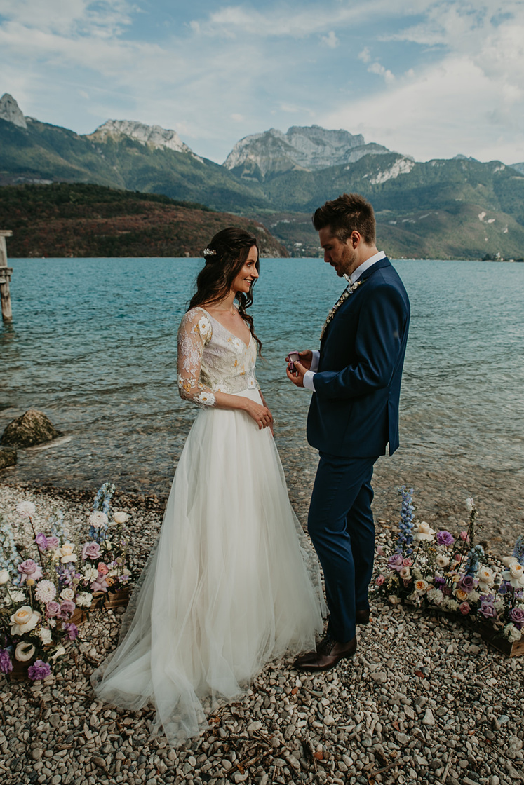 Couple de mariés en train de se passer la bague au doigt lors d'une cérémonie laïque au bord du lac d'Annecy. 
La mariée porte une robe blanche en tulle avec un haut en broderie blanche et jaune pastel à manches. Le marié porte un costume bleu marine